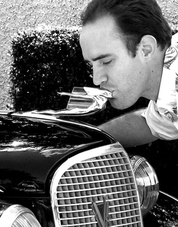 Black and white photograph of a man leaning down to kiss the chrome hood ornament of a classic black car.