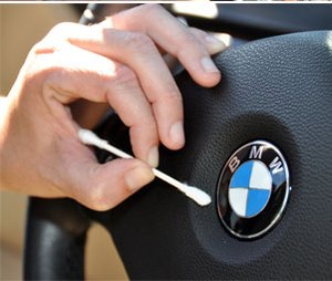 A hand using a cotton swab to clean around a car’s steering wheel badge.