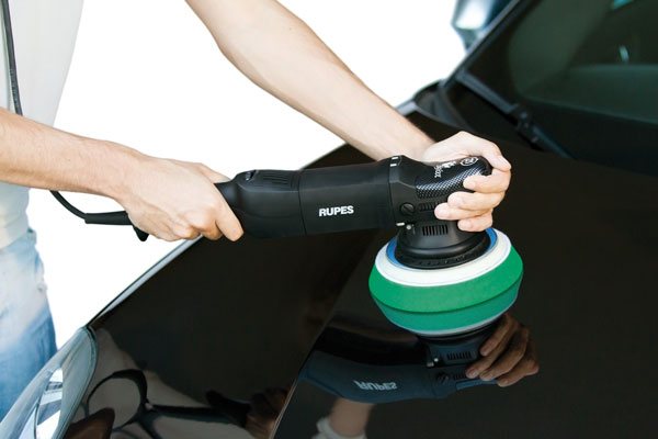 A dual-action polisher with a green pad being worked slowly over a black car panel.