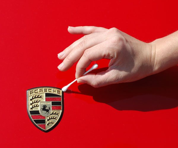 A hand using a cotton swab to clean around the raised crest badge on a red car&rsquo;s bonnet.
